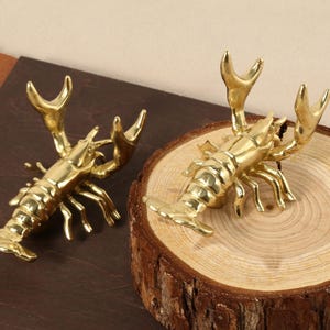May include: Two gold-coloured lobster sculptures on a wooden surface. The lobsters are detailed and realistic, with claws and antennae.