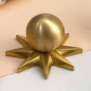 Könnte beinhalten: Ein goldfarbener Möbelknopf aus Metall mit einer kugelförmigen Oberseite, die auf einer flachen, sternförmigen Basis mit zwölf spitzen Strahlen zentriert ist. Die Metalloberfläche hat ein gebürstetes, mattes Finish für ein einheitliches Aussehen.