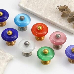 Può includere: Una collezione di otto pomelli rotondi per armadietti in vari colori, tra cui blu scuro, azzurro, arancione, rosa, bianco, verde e viola. Ogni pomello presenta una parte superiore circolare liscia e traslucida con un piccolo perno metallico.