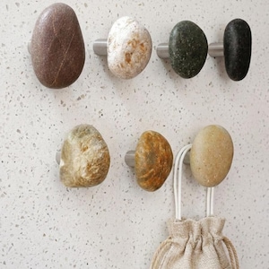May include: A set of seven wall-mounted hooks, each featuring a unique natural river stone as the decorative knob. The stones vary in color, including shades of brown, white, grey, green, and tan, and are attached to small, cylindrical metal bases.