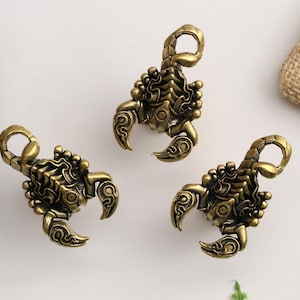 May include: Three antique brass-toned metal scorpion charms, each featuring a detailed exoskeleton texture, curved pincers, and a raised tail loop for attachment. The metallic finish displays dark highlights in the recessed areas.
