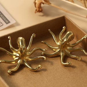 Op de afbeelding: Twee gouden octopus-beeldjes op een bruin oppervlak. De octopus-beeldjes zijn gedetailleerd en hebben een realistisch uiterlijk.