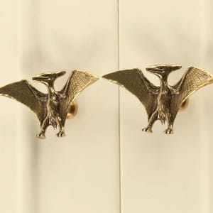 Peut inclure: Deux boutons de cabinet en forme de ptérodactyles aux ailes déployées, de couleur dorée.