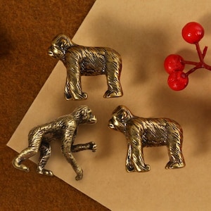 May include: Three bronze-colored monkey figurines in different poses are displayed on a brown and tan surface. A sprig of red berries adds a pop of color. The figurines have detailed textures and are likely decorative items.