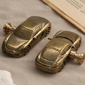 May include: Two antique brass-toned metal cabinet knobs shaped like sports cars. Each knob features a detailed car body with a smooth finish and a cylindrical mounting post extending from the side, designed for attachment to furniture or cabinetry.