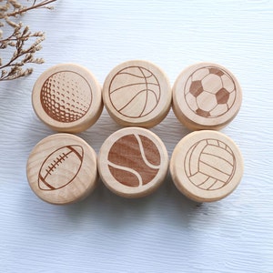 May include: Six wooden cabinet knobs with engraved sports ball designs: golf, basketball, soccer, football, tennis, and volleyball. The knobs are light-colored wood with dark brown etched details, arranged in two rows of three.