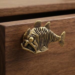 Puede incluir: Un tirador de mueble de metal color latón con forma de pez de aguas profundas, con un ojo prominente, dientes afilados y aletas texturizadas. El mango con forma de pez está montado en el frente de un cajón de madera de color marrón oscuro.