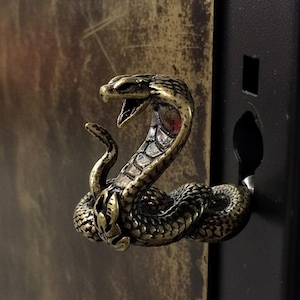 May include: A bronze-coloured cobra-shaped cabinet pull. The snake is coiled with its hood flared and mouth open, revealing a red interior. The pull is attached to a black cabinet door, with a textured, aged background.