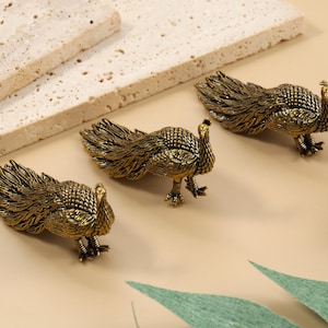 May include: Three antique gold peacock figurines with detailed feather designs. The peacocks are positioned on a light beige surface, with two rectangular stone blocks in the background. The figurines are a decorative accent.