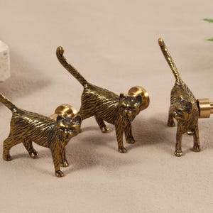 May include: Three brass cat-shaped cabinet knobs. The knobs are detailed with textured fur and stand on small feet. The cat knobs are in a standing pose with their tails up. The knobs are a warm gold colour.