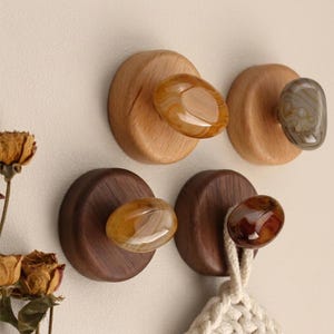 May include: Four circular wall hooks featuring polished, smooth, oval-shaped stones in shades of amber, brown, and grey, mounted on round wooden bases. Two hooks are made of light-colored wood, and two are made of dark-colored wood.