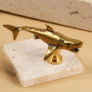 May include: A golden shark-shaped cabinet knob mounted on a square, off-white stone. The shark has detailed fins, gills, and an open mouth. The knob is made of brass and is a decorative hardware item.