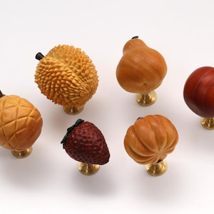 Puede incluir: Un conjunto de seis pomos para armarios con forma de fruta que incluye una piña, un durián, una pera, una manzana roja, una fresa y una calabaza. Cada pomo presenta texturas y colores realistas sobre una pequeña base de tono latón.