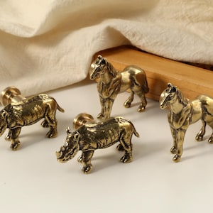 Puede incluir: Cuatro tiradores de armario de latón con forma de animales. Dos son rinocerontes y dos son caballos. Los tiradores tienen detalles realistas y un acabado cálido y antiguo. Se colocan sobre un fondo neutro con una bandeja de madera y un paño color crema.