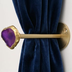 May include: A wall-mounted curtain tieback featuring a gold-toned metal rod and circular base. The end of the rod is capped with a polished, irregular purple agate slice that has a textured, gold-colored metallic edge, holding a navy blue velvet curtain.