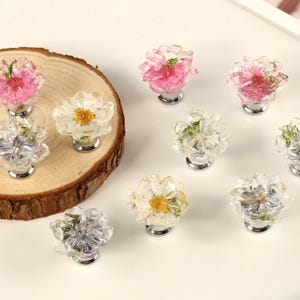 以下が含まれることがあります： 透明な樹脂とシルバーベースのフローラルキャビネットノブのコレクション。ノブには、ピンク、白、紫の色合いの本物の花が使用されています。木のスライスに配置されたノブもあれば、白い表面にあるノブもあります。