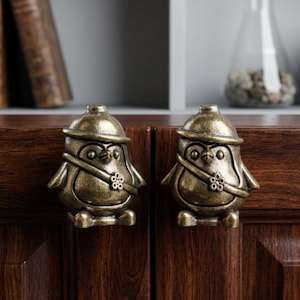 Peut inclure: Deux boutons de placard en métal couleur laiton antique en forme de pingouins. Chaque figurine de pingouin porte un chapeau et une sangle diagonale avec un petit détail en forme de fleur. Les boutons ont une finition texturée et vieillie.