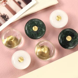 May include: Six decorative drawer pulls with a gold accent. The pulls are made of different materials, including green marble, clear glass, and white marble.