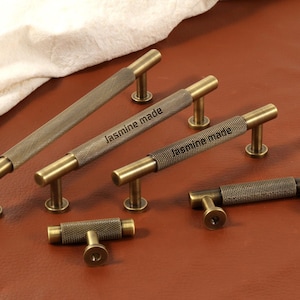 May include: A selection of antique brass cabinet pulls and handles. The handles feature a textured, diamond-patterned grip and are labelled "Jasmine made". The hardware is in various sizes and shapes, with a warm, aged finish.