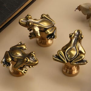 Op de afbeelding: Drie messing kikkerbeeldjes met gedetailleerde kenmerken, elk gemonteerd op een kleine, ronde basis. De kikkers staan in verschillende poses, met een warme, antieke afwerking. De achtergrond is een neutraal beige.