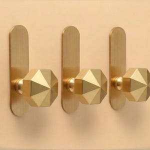 May include: Three wall-mounted hooks made of brushed brass. Each hook features a vertical rectangular backplate with rounded ends and a protruding, faceted, octagonal knob. The metal has a consistent matte gold-toned finish throughout.