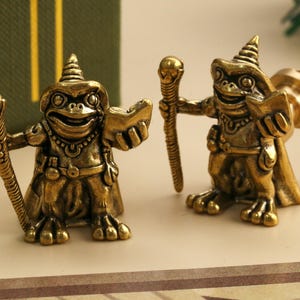 May include: Two brass frog figurines, each wearing a wizard hat and cape, holding a staff and a book. The figurines have detailed features and a vintage aesthetic. The background includes a green box with gold lettering.