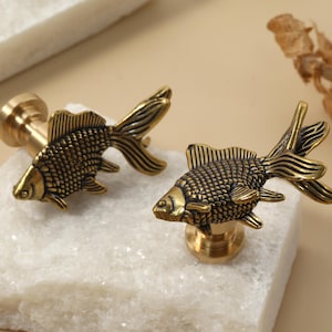 May include: Two antique brass goldfish cabinet knobs. The knobs are detailed with scales and fins, and are mounted on a brass base. The knobs are displayed on a white stone surface.