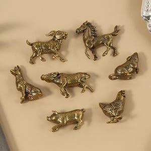 Op de afbeelding: Een verzameling kleine, bronskleurige dierenbeeldjes, waaronder een paard, een geit, een hond, een koe, een varken, een rat en een kip. De beeldjes zijn gedetailleerd en lijken van metaal te zijn. Boerderij- of dierenriemthema.