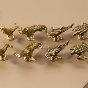May include: A collection of eight brass animal-shaped cabinet knobs. The set includes knobs shaped like seals, sharks, and turtles. Each knob is detailed with textured surfaces and mounted on a small, round base. The knobs are in a warm, golden color.