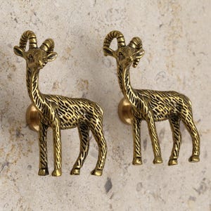 May include: Two brass-toned cabinet knobs shaped like stylized deer. Each knob features a deer with curved horns, a long neck, and detailed body texture. The knobs are mounted on round, brass-colored bases.