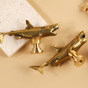 May include: Two brass-toned metal cabinet knobs shaped like sharks. Each shark figure features an open mouth with visible teeth, a dorsal fin, and a tail. The knobs have a cylindrical base with a threaded hole for attachment to furniture.