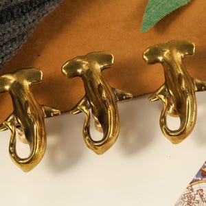 May include: Three gold-toned hammerhead shark shaped hooks. The hooks are mounted on a brown surface.