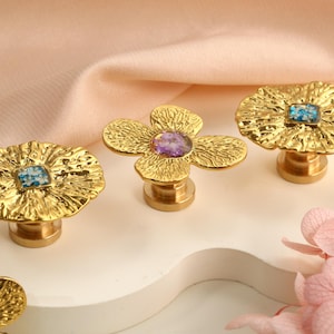 May include: Close-up of three gold-colored flower-shaped cabinet knobs with textured surfaces. Each knob features a gemstone center, with blue and purple stones. The knobs are on a white surface with a pink background.