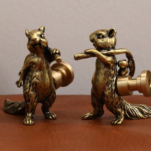 May include: Two bronze squirrel figurines, each holding a cylindrical object. The squirrels have detailed fur and are posed in a standing position. The background is a neutral color, highlighting the metallic finish of the figurines.