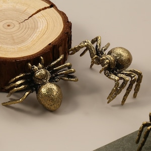 May include: Three bronze-coloured spider figurines with detailed legs and bodies are displayed. The spiders are positioned on a light surface near a wooden slice and a piece of wood. The spiders are a decorative item.