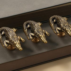 Op de afbeelding: Drie messing kastknoppen in de vorm van een alligator. De knoppen hebben gedetailleerde schubtexturen en zijn gemonteerd op ronde bases. De kop en staart van de alligator vormen een handvat. De knoppen liggen op een donkergrijze ondergrond.