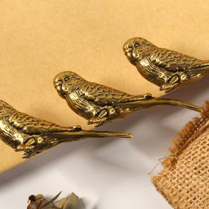 Op de afbeelding: Drie antieke goudkleurige decoratieve clips in de vorm van vogels. De vogels zitten op takken, met gedetailleerde veren en oogkenmerken. De clips liggen op een lichtbruin oppervlak, met een jute zak op de achtergrond.