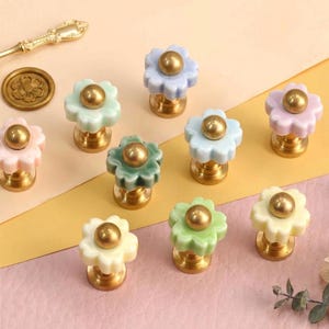 May include: A collection of nine flower-shaped wax seal stamps with pastel-colored ceramic petals and brass centers, accompanied by a small gold-toned metal spoon and a circular brass seal disc featuring a floral engraving on the top surface.