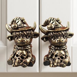 May include: Two bronze-toned cabinet knobs shaped like Highland cows wearing witch hats with small pumpkins on the brims. Each cow figure has long, textured fur, curved horns, and is holding a human skull in its front paws, creating a detailed design.