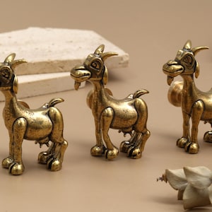 Może przedstawiać: Trzy antyczne, złote figurki kóz. Każda koza ma szczegółowy design, w tym rogi, brodę i uśmiechniętą ekspresję. Kozy stoją na jasnobrązowej powierzchni, z neutralnym tłem. Figurki są prawdopodobnie przedmiotami dekoracyjnymi.