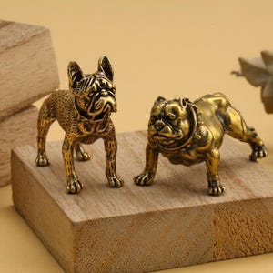 May include: Two brass bulldog figurines, one standing and one crouching, are displayed on a wooden block. The dogs have detailed features and a golden finish. The background is a neutral color, highlighting the figurines.