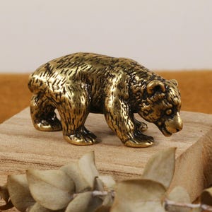May include: A small, detailed brass bear figurine. The bear is posed in a walking stance, with textured fur and a slightly lowered head. It is placed on a small wooden block, with other bear figurines visible.