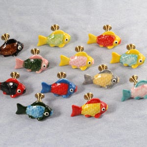 Puede incluir: Surtido de 12 tiradores de cajones de cerámica con forma de pez de colores. Cada tirador presenta un color y un diseño diferente, incluyendo rojo, azul, verde, amarillo y negro. Los peces están todos mirando hacia la derecha.