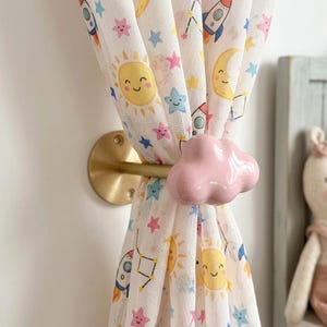 Può includere: Un fermatenda con una nuvola in ceramica rosa chiaro lucido fissata a un supporto a parete in metallo dorato spazzolato. Il fermatenda trattiene una tenda in tessuto bianco stampata con un motivo colorato di soli, lune, razzi e stelle sorridenti.