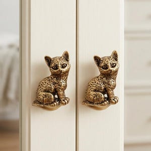 May include: Two identical brass-toned metal cabinet knobs, each shaped like a small sitting cat with detailed fur texture and pointed ears. The knobs are mounted on a vertical white wooden surface, serving as functional handles for cabinet doors.