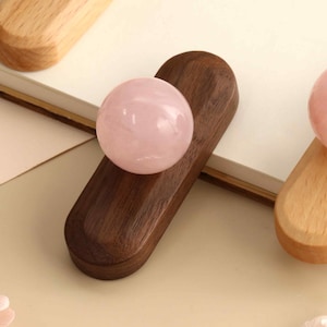 May include: Three smooth, polished, light pink spherical stones, each resting on an elongated, rounded wooden base. One base is dark brown wood, while the other two are light-colored wood, all featuring a central groove to hold the sphere.