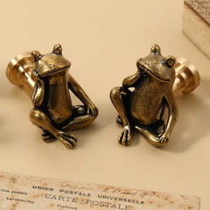 Może przedstawiać: Dwie antyczne pieczęcie woskowe w kształcie żaby z mosiądzu. Żaby siedzą, z jedną ręką na brodzie. Pieczęcie mają mosiężny uchwyt. Na pierwszym planie znajduje się stara pocztówka.