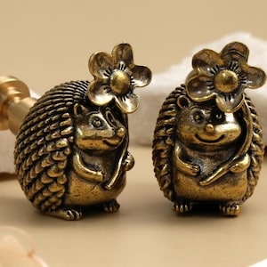 May include: Two antique brass hedgehog figurines with flower accents. The hedgehogs have detailed spines and smiling faces. One figurine is attached to a brass stem. Decorative items for home decor.