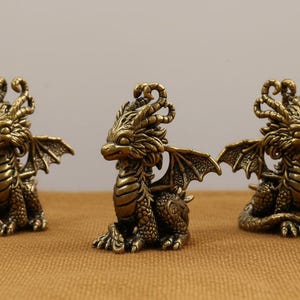 Op de afbeelding: Drie bronzen drakenbeeldjes. Elke draak zit met gedetailleerde vleugels, schubben en een decoratieve kop. De draken worden tentoongesteld op een bruinachtige ondergrond.