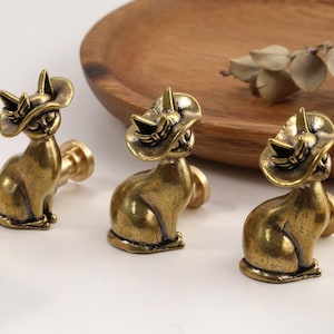 May include: Three brass-toned metal cabinet knobs, each shaped like a seated cat wearing a wide-brimmed hat. The figures have a polished, antique-style finish with visible dark detailing in the crevices, and each features a cylindrical base.
